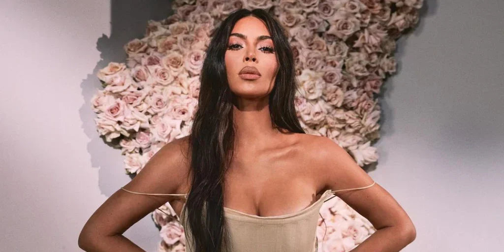 Kim Kardashian in a beige dress, standing hands-on-hips in front of a floral wall of roses, with long, sleek dark hair.